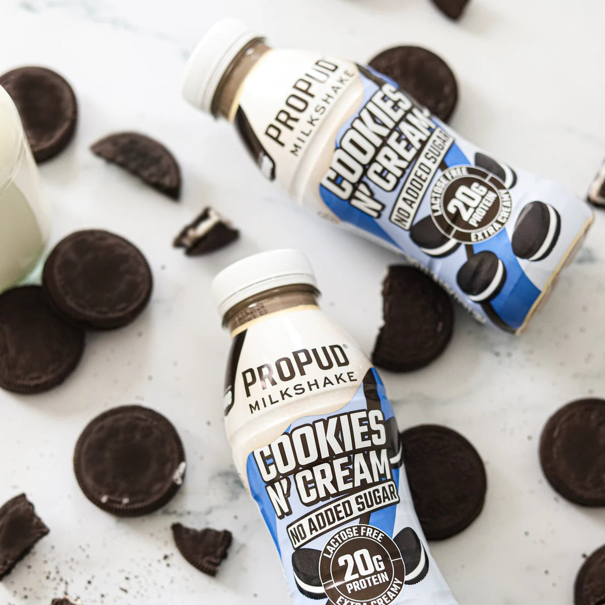 Two bottles of Propud Milkshake Cookies N' Cream protein drink arranged on a white marble surface with Oreo cookies and cookie crumbs around them.